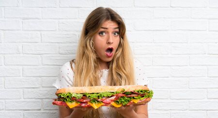 Young blonde woman holding a big sandwich making surprise gestureの写真素材