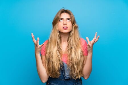 Young blonde woman with overalls over isolated blue wall frustrated by a bad situation