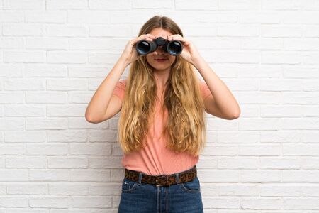 Young blonde woman over white brick wall with black binocularsの写真素材