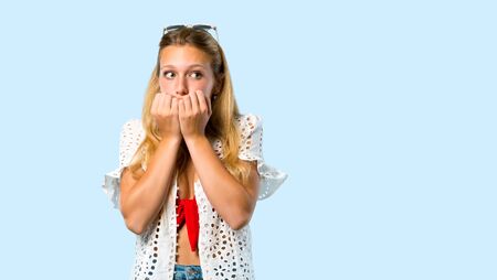 Blonde girl in summer vacation is a little bit nervous and scared putting hands to mouth on blue backgroundの写真素材