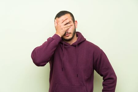 Colombian man with sweatshirt over green wall covering eyes by handsの写真素材