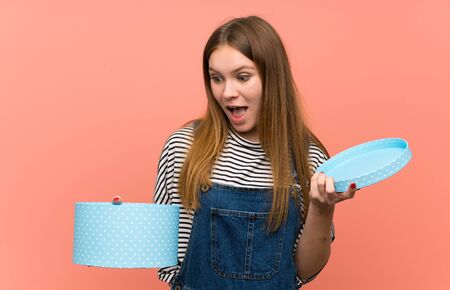 Young woman with overalls over pink wall holding a gift in hands