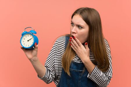 Young woman with overalls over pink wall holding vintage alarm clock
