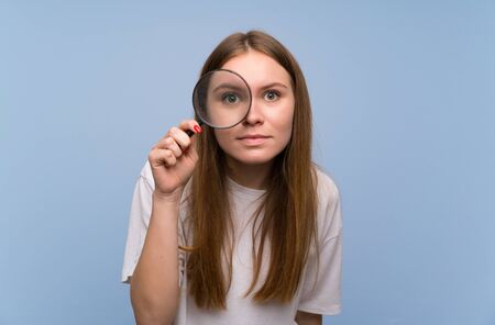 Young woman over blue wall taking a magnifying glass and looking through itの販売画像