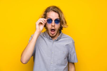 Blonde man over isolated yellow background with glasses and surprisedの写真素材