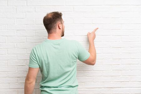 Blonde man over brick wall pointing back with the index fingerの写真素材