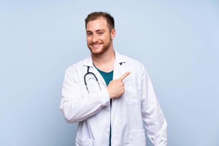 Handsome doctor man over blue wall pointing to the side to present a productの写真素材