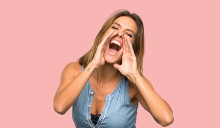 Blonde woman with jean dress shouting and announcing something over isolated pink background