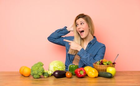 Young blonde woman with many vegetables surprised and pointing finger to the sideの写真素材