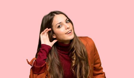 Young woman with coat listening to something by putting hand on the ear over isolated pink backgroundの写真素材