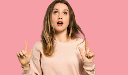 Teenager girl with pink sweater surprised and pointing up over isolated pink backgroundの写真素材