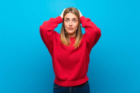 Woman with red sweater over blue wall frustrated and takes hands on headの写真素材