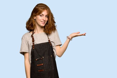 Young redhead woman with apron holding copyspace imaginary on the palm to insert an ad over blue backgroundの写真素材