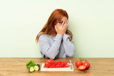 Teenager redhead girl with vegetables in a table covering eyes and looking through fingersの写真素材