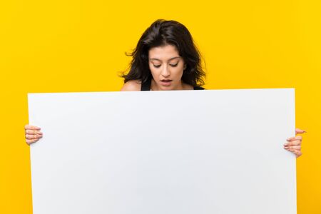 Young woman over isolated yellow background holding an empty white placard for insert a conceptの写真素材