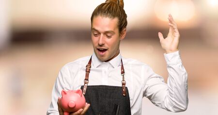 Barber man in an apron surprised while holding a piggybank in a barber shopの写真素材