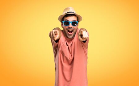 Man with hat and sunglasses on his summer vacation points finger at you while smiling on orange backgroundの写真素材