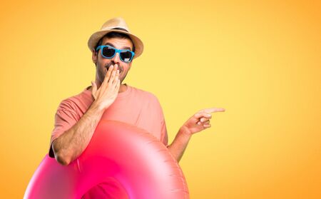 Man with hat and sunglasses on his summer vacation pointing finger to the side and presenting a product with a surprised face while covering mouth on orange backgroundの写真素材