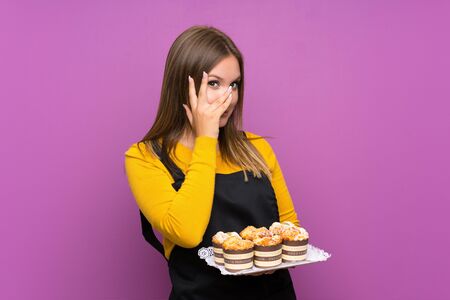 Teenager girl holding lots of different mini cakes over isolated purple background covering eyes and looking through fingersの写真素材