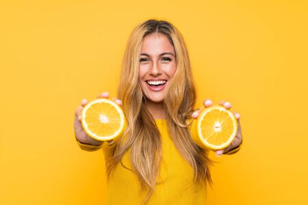 Young blonde woman holding an orange over yellow backgroundの写真素材