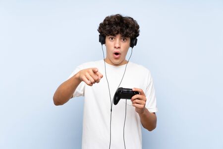 Young man playing with a video game controller over isolated blue wall surprised and pointing frontの写真素材