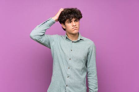 Young man over isolated purple wall having doubts while scratching headの写真素材