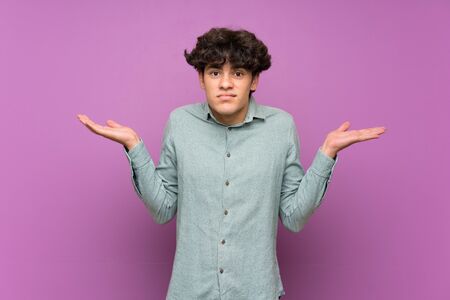 Young man over isolated purple wall having doubts while raising handsの写真素材