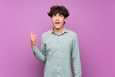 Young man over isolated purple wall pointing up and surprisedの写真素材