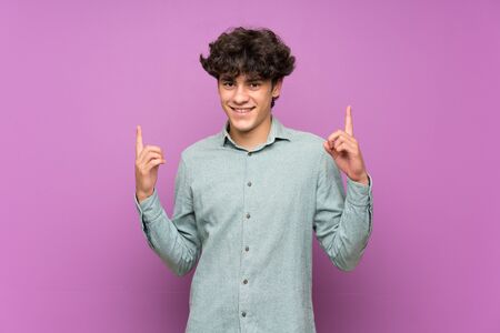 Young man over isolated purple wall pointing up a great ideaの写真素材