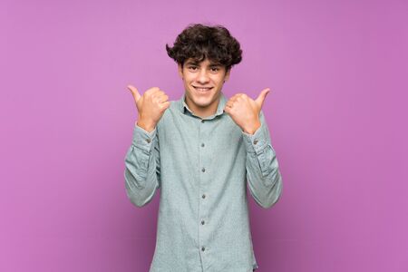 Young man over isolated purple wall with thumbs up gesture and smilingの写真素材