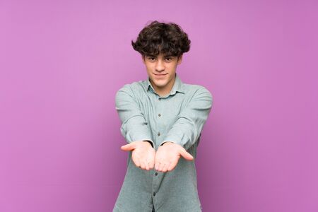 Young man over isolated purple wall holding copyspace imaginary on the palm to insert an adの写真素材