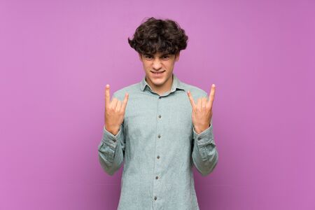 Young man over isolated purple wall making rock gestureの写真素材