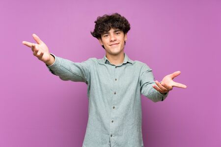 Young man over isolated purple wall presenting and inviting to come with handの写真素材