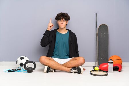 Young sport man sitting on the floor around many sport elements pointing with the index finger a great ideaの写真素材