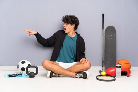 Young sport man sitting on the floor around many sport elements pointing finger to the sideの写真素材