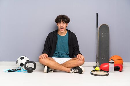 Young sport man sitting on the floor around many sport elements with surprise facial expressionの写真素材