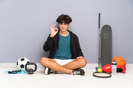 Young sport man sitting on the floor around many sport elements showing an ok sign with fingersの写真素材