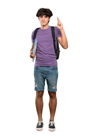 A full-length shot of a Young student man with fingers crossing and wishing the best over isolated white backgroundの写真素材