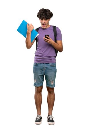 A full-length shot of a Young student man with phone in victory position over isolated white backgroundの写真素材