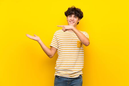 Young man over isolated yellow wall holding copyspace imaginary on the palm to insert an adの写真素材