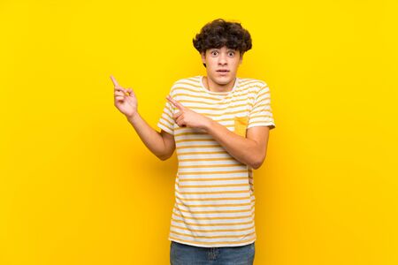 Young man over isolated yellow wall frightened and pointing to the sideの写真素材