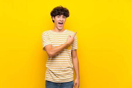 Young man over isolated yellow wall surprised and pointing sideの写真素材