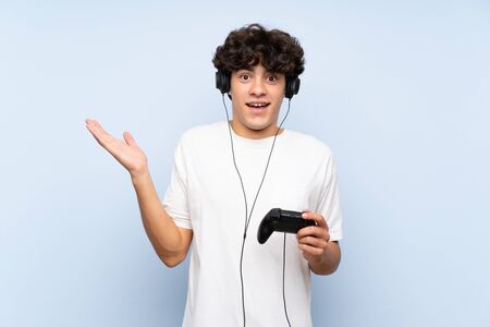 Young man playing with a video game controller over isolated blue wall with shocked facial expressionの写真素材