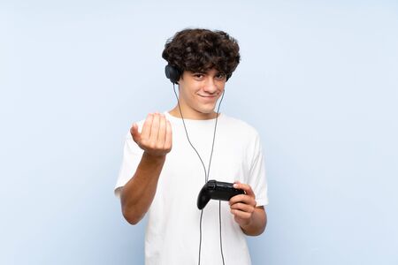 Young man playing with a video game controller over isolated blue wall inviting to come with hand. Happy that you cameの写真素材