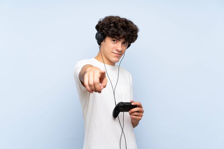 Young man playing with a video game controller over isolated blue wall points finger at you with a confident expressionの写真素材
