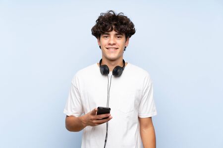 Young man listening music with a mobile over isolated blue wall smiling a lotの写真素材