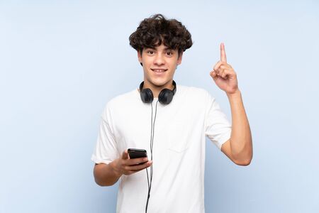 Young man listening music with a mobile over isolated blue wall pointing up a great ideaの写真素材