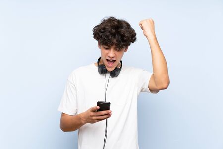 Young man listening music with a mobile over isolated blue wall celebrating a victoryの写真素材