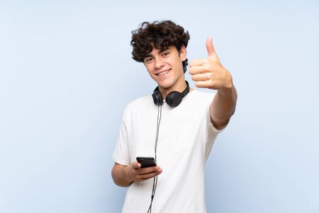 Young man listening music with a mobile over isolated blue wall with thumbs up because something good has happenedの写真素材