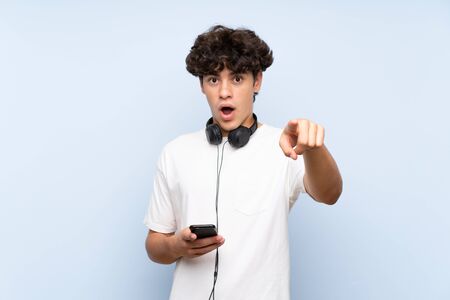 Young man listening music with a mobile over isolated blue wall surprised and pointing frontの写真素材